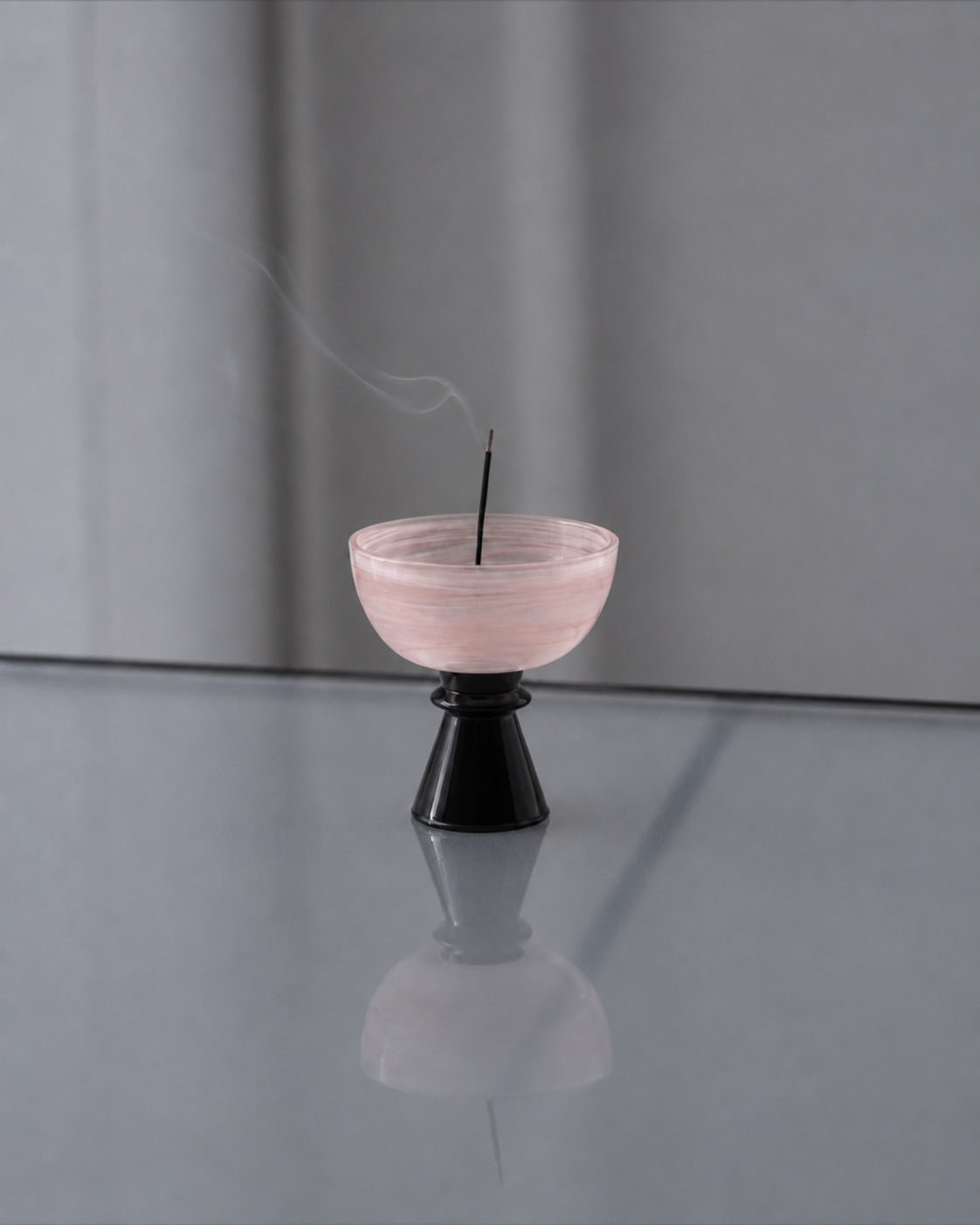 Rosé Cloud Glass Incense Holder burning stick incense on a reflective gray surface, with delicate smoke rising in a quiet minimalist interior.