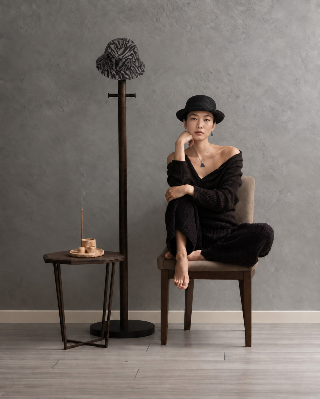 Woman in black seated beside burning incense, sculptural ceramic holders, and hats in a quiet minimalist interior.