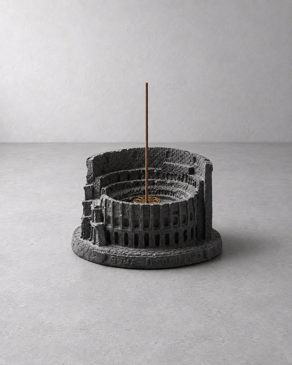 A dark gray textured resin incense holder shaped like a classical amphitheater, with a single incense stick standing vertically in the center, set against a minimalist gray concrete background.