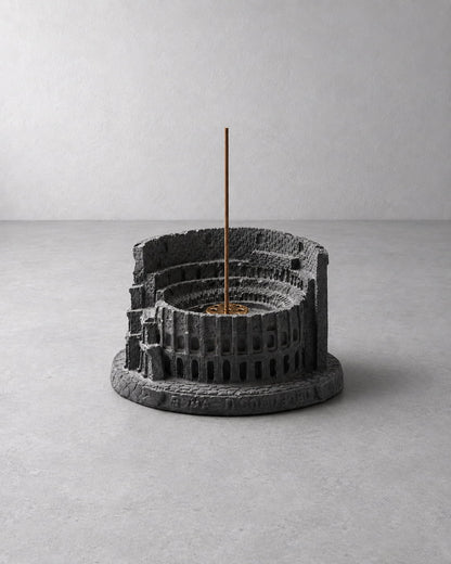 A dark gray textured resin incense holder shaped like a classical amphitheater, with a single incense stick standing vertically in the center, set against a minimalist gray concrete background.