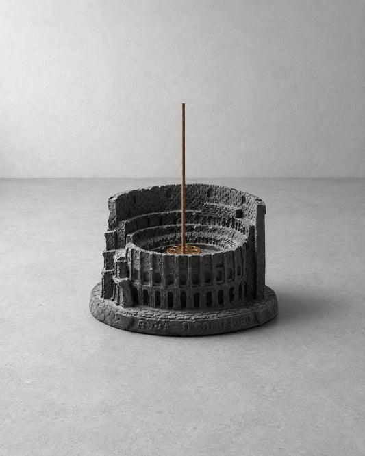 A dark gray textured resin incense holder shaped like a classical amphitheater, with a single incense stick standing vertically in the center, set against a minimalist gray concrete background.