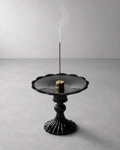 A black ceramic incense burner with a fluted pedestal base and scalloped rim, holding a smoking incense stick on a light gray background.