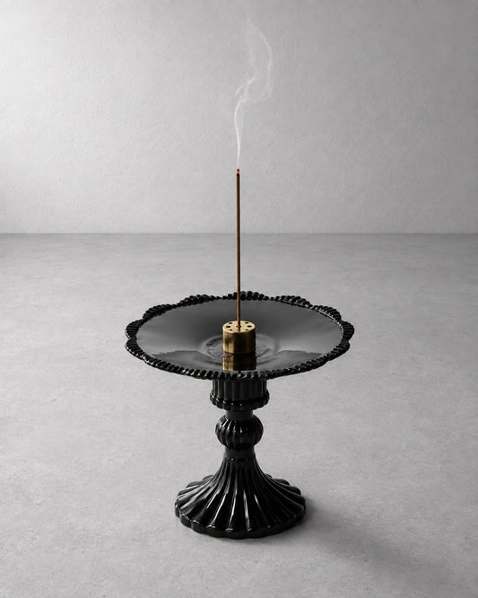 A black ceramic incense burner with a fluted pedestal base and scalloped rim, holding a smoking incense stick on a light gray background.