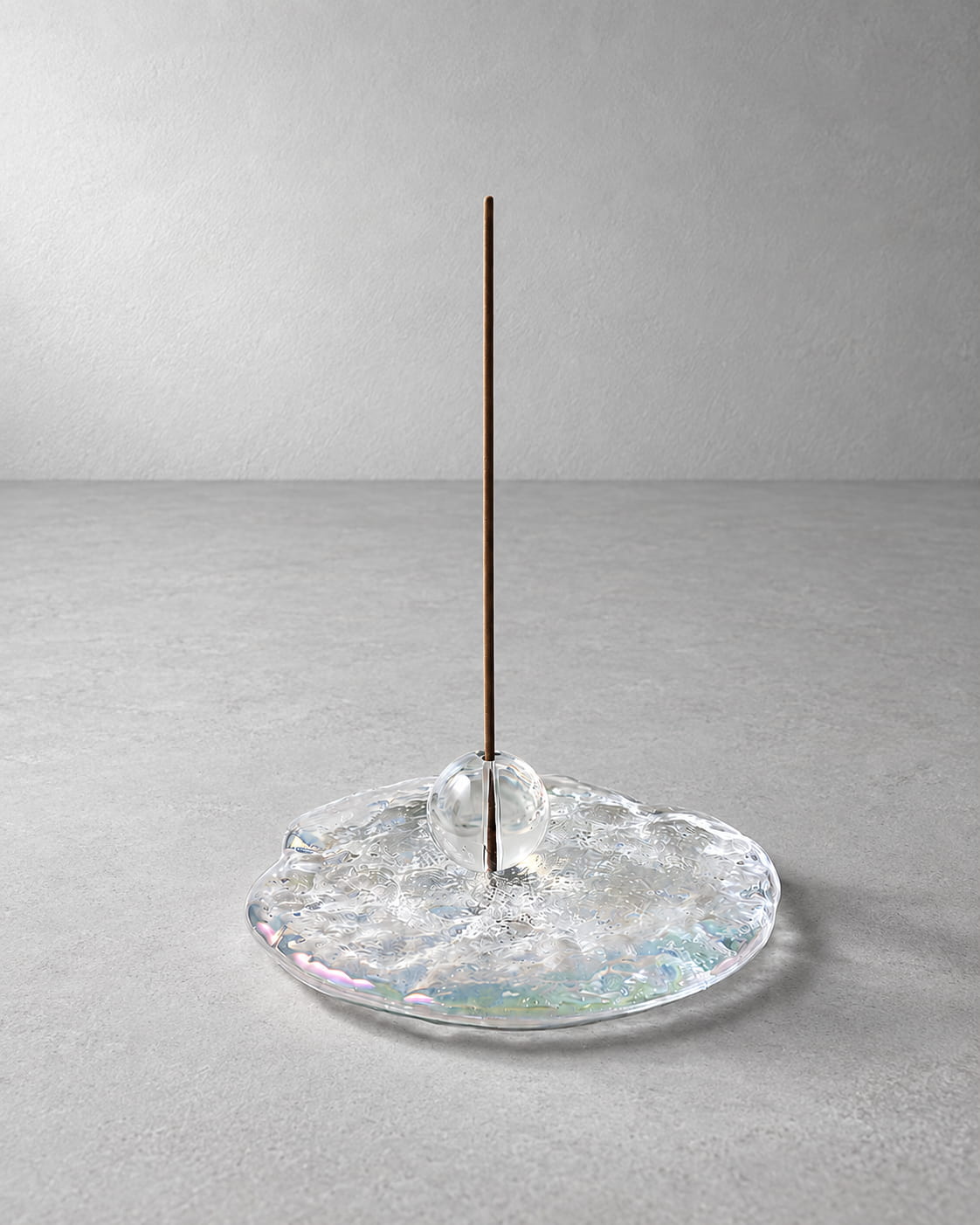 A minimalist incense holder featuring a single vertical brown incense stick standing in a clear, textured glass base with subtle iridescent reflections, set against a neutral gray background.