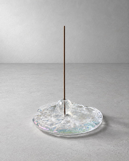 A minimalist incense holder featuring a single vertical brown incense stick standing in a clear, textured glass base with subtle iridescent reflections, set against a neutral gray background.