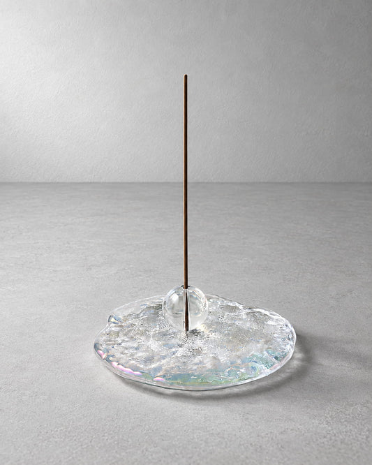 A minimalist incense holder featuring a single vertical brown incense stick standing in a clear, textured glass base with subtle iridescent reflections, set against a neutral gray background.