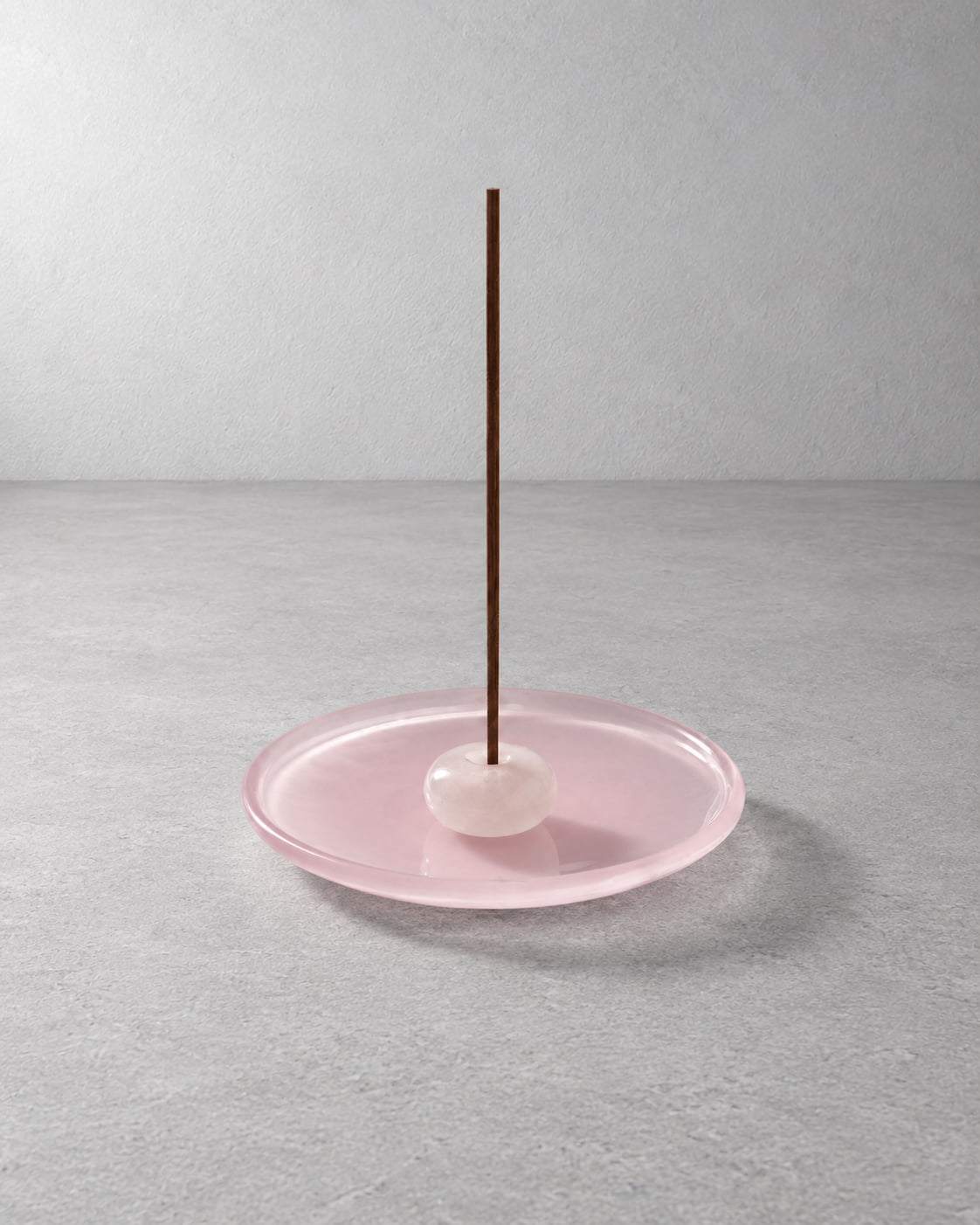 A minimalist incense holder featuring a round translucent pink glass dish, a smooth pale pink stone ball, and a single dark brown incense stick standing vertically, set against a neutral gray textured background.